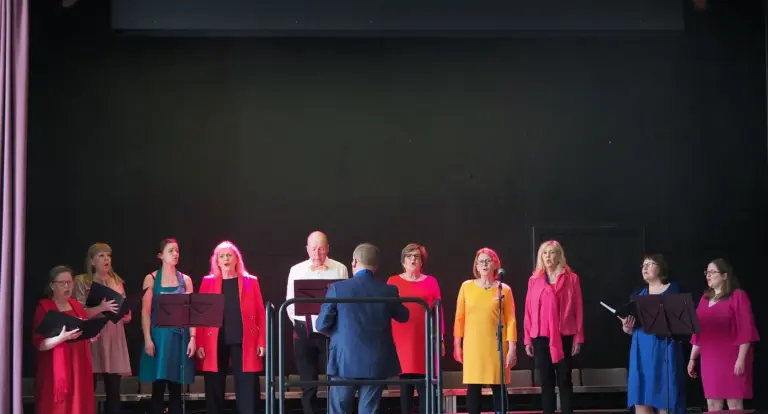 Värikkäästi pukeutuneita kuorolaulajia seisoo esiintymislavalla.